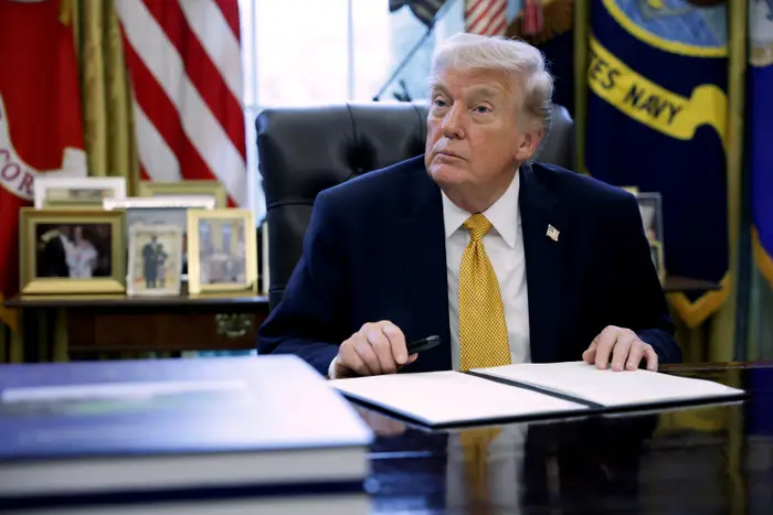 En person i dress sitter ved et skrivebord i offisielle omgivelser med flagg og innrammede bilder i bakgrunnen, og holder en penn over et dokument