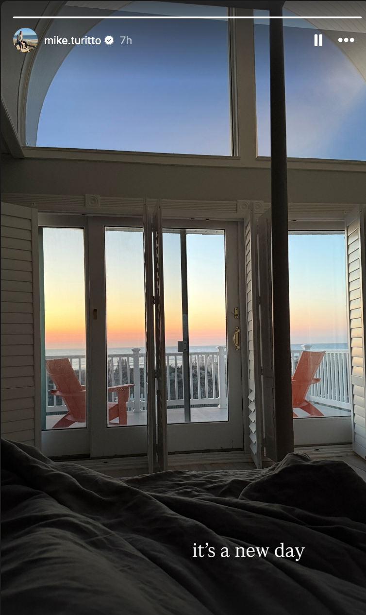 Utsikt fra en seng av en soloppgang gjennom doble dører, med to stoler på en balkong. Teksten lyder 