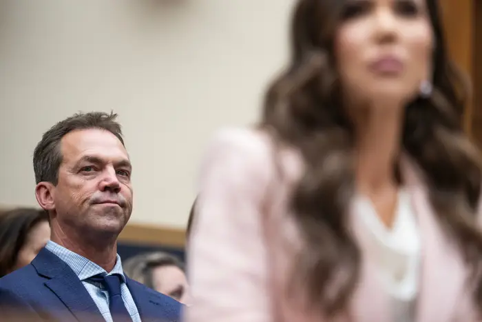 En mann i dress lytter oppmerksomt i en formell setting, mens en kvinne i forretningsantrekk er i forgrunnen