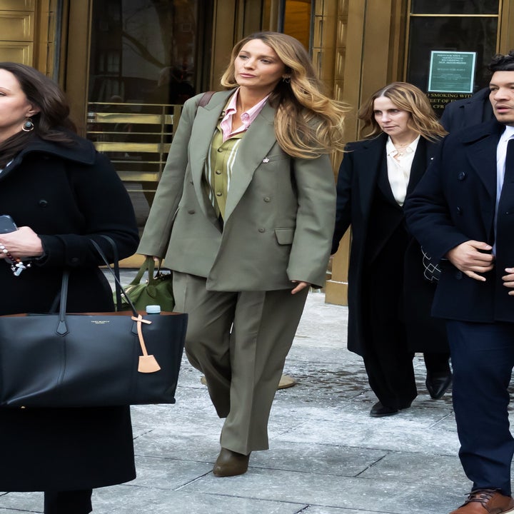 Person i en skreddersydd dress, bluse og vest går utendørs, flankert av andre i frakker, dukker opp på et offentlig arrangement eller sammenkomst