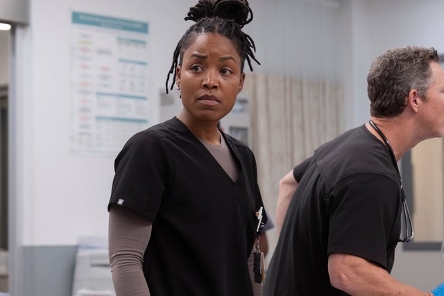 Medisinsk dramascene med en sykepleier i svart skrubb, ser alvorlig og fokusert ut. En annen person er ved siden av henne, delvis vendt bort