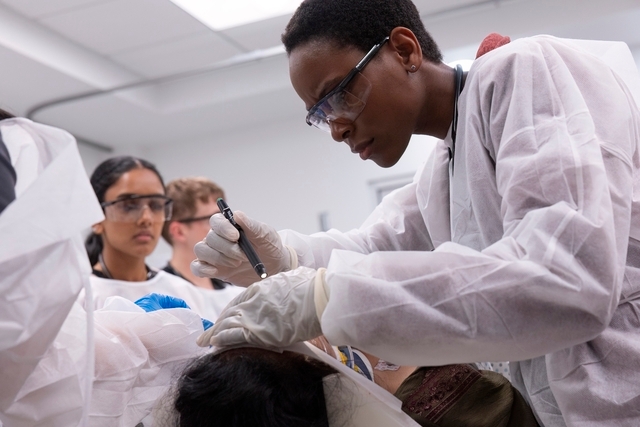 Person i laboratoriefrakk og vernebriller som utfører en prosedyre mens andre observerer i et klinisk miljø