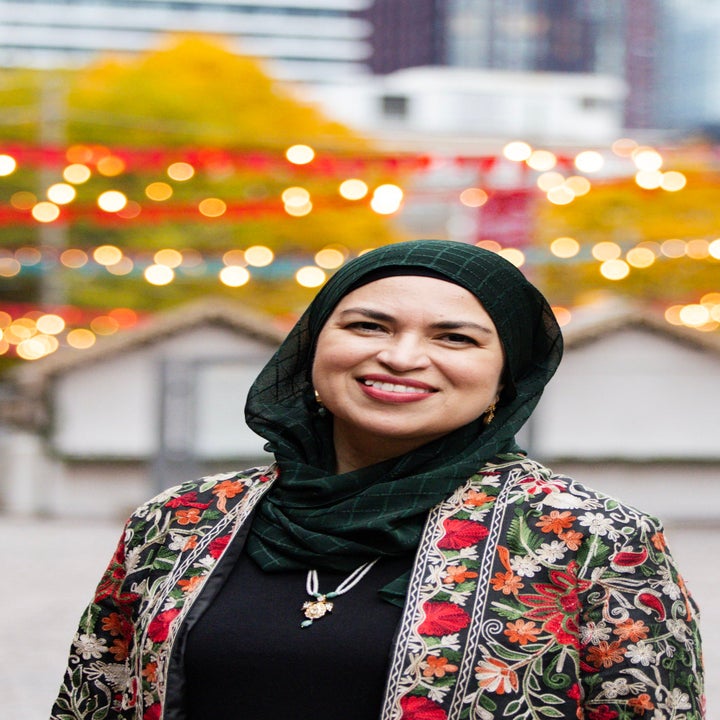 Person som smiler, iført en mønstret jakke og hodeskjerf, i en utendørs setting med uskarpe lys og bygninger i bakgrunnen