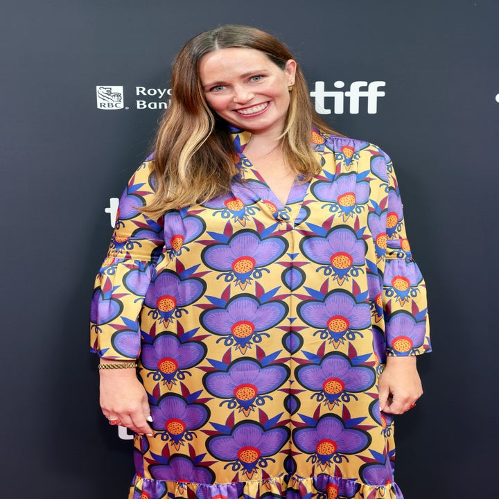 Person som smiler i dristig blomsterkjole på en filmbegivenhet; stående mot et bakteppe med sponsorlogoer