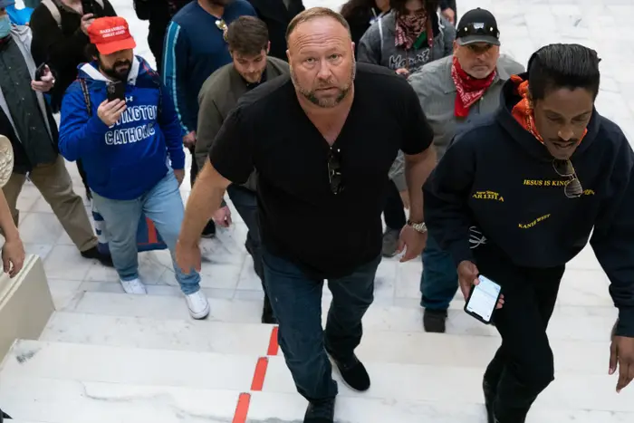 En gruppe mennesker, noen med "Trump" hatter og skjorter, gå opp trapper i en bygning. En mann foran holder en telefon. Kameraer og trykk er i bakgrunnen