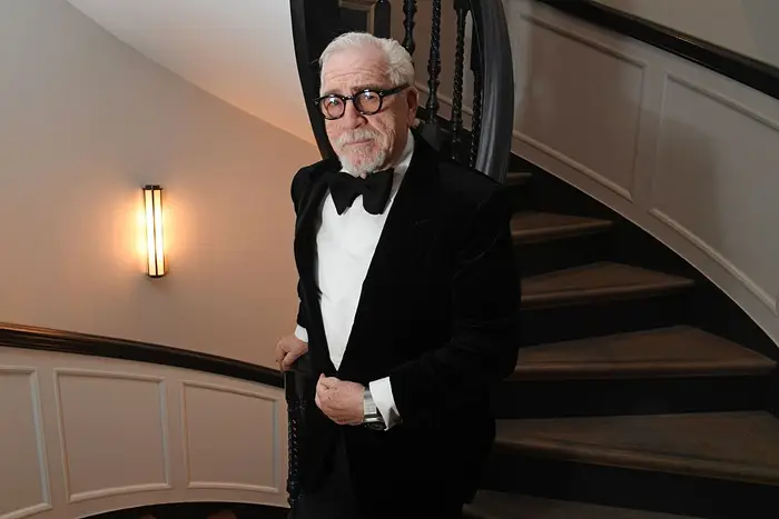 En eldre mann i formell antrekk, med briller og smoking, står på en buet trapp og ser selvsikkert på kameraet