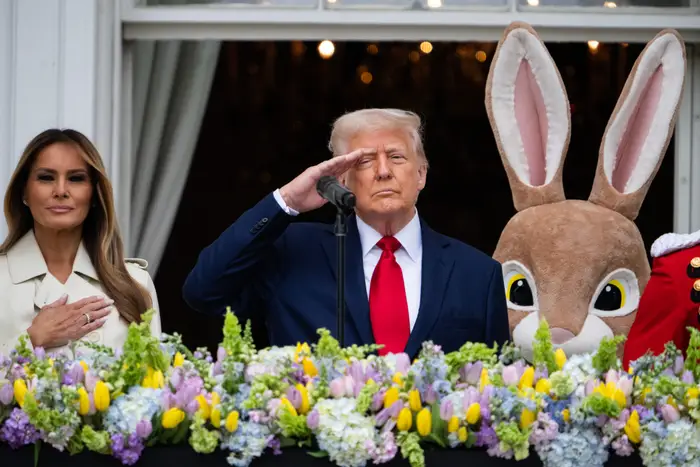 En mann i dress hilser på en blomsterdekorert balkong ved siden av en kvinne og en person i kanindrakt