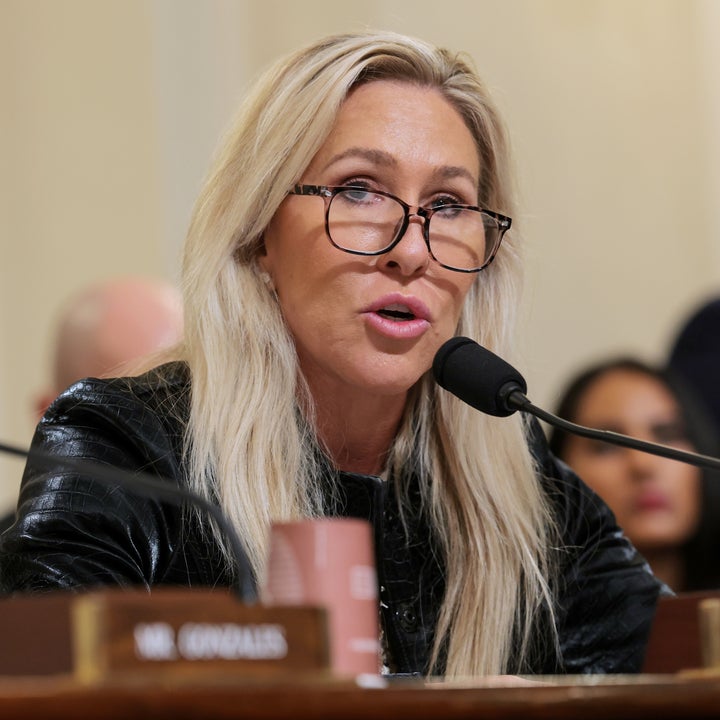 Person som snakker i en mikrofon på et møte, iført briller og en skinnlignende jakke, med folk som sitter i bakgrunnen