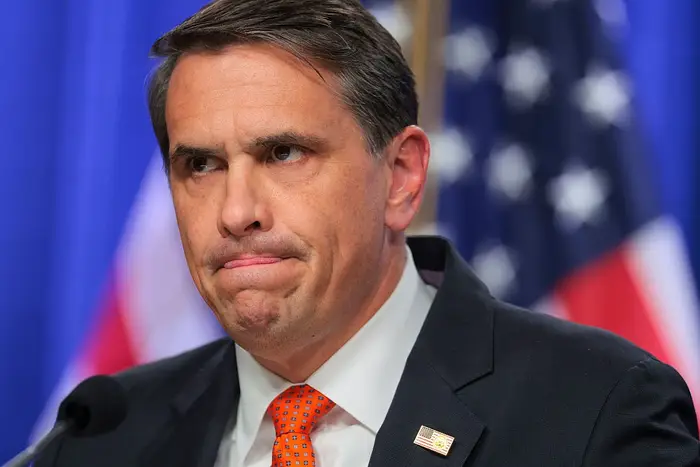 Mann i dress med en amerikansk flaggnål står foran et amerikansk flagg i bakgrunnen og virker gjennomtenkt eller bekymret