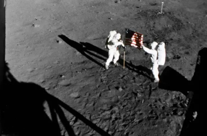 Astronauter i romdrakter står på måneoverflaten, en holder et flagg som symboliserer den historiske månelandingen