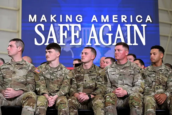 Fem soldater i uniform sitter foran et skilt med teksten «Making America Safe Again»; under et arrangement