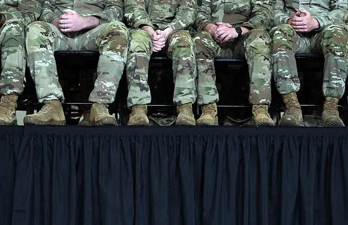 En rekke mennesker i kamuflasjeuniformer og støvler sitter på en scene med hendene sammen, bena synlige under et svart gardin