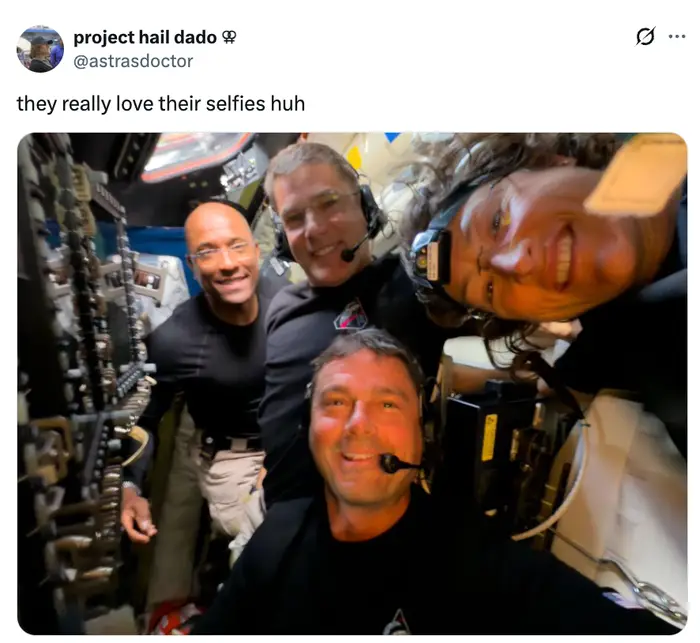 Fire personer i en romfartøys cockpit, smiler og tar en selfie. De ser ut til å være astronauter eller i et simuleringsmiljø