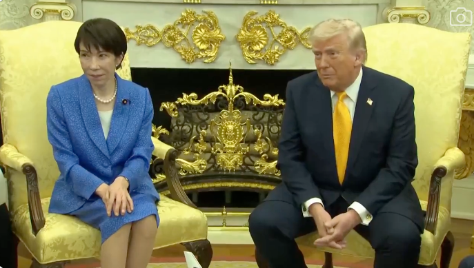 To personer sitter i en formell setting. Personen til venstre har på seg en blå dress, mens personen til høyre har på seg en dress med gult slips