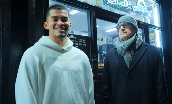 To personer som står utenfor en kafé. Man har på seg hettegenser og smiler; den andre har på seg frakk, skjerf og hatt og ser seriøs ut