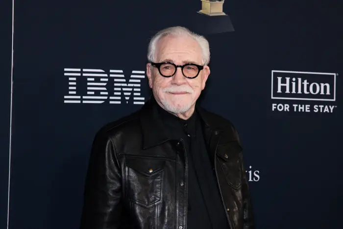 En eldre mann med hvitt hår og briller iført en svart skinnjakke poserer på et formelt arrangement. Bakgrunnen har IBM- og Hilton-logoer