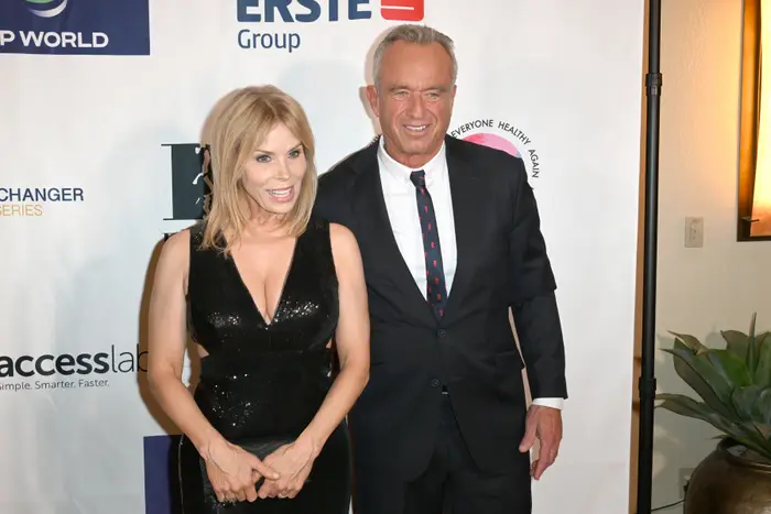 Cheryl Hines og RFK Jr. smiler på en rød løper. Kvinnen har på seg en ermeløs svart kjole, og mannen er i dress med mønstret slips