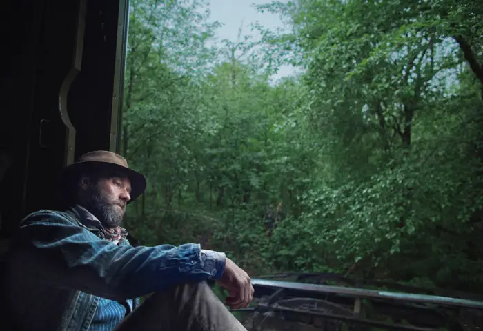 Mann i lue og dongerijakke sitter ettertenksomt ved et åpent vindu, med utsikt over en frodig, grønn skog
