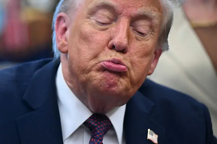 En person med lukkede øyne som lager et buldrende ansiktsuttrykk, iført en dress med en amerikansk flagg jakkeslagsnål