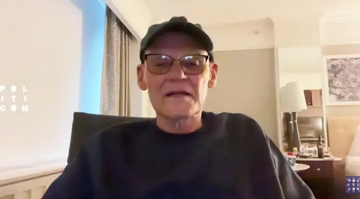 En person med briller og caps sitter i et rom med lyse vegger, synlig bak dem er en seng og lampe