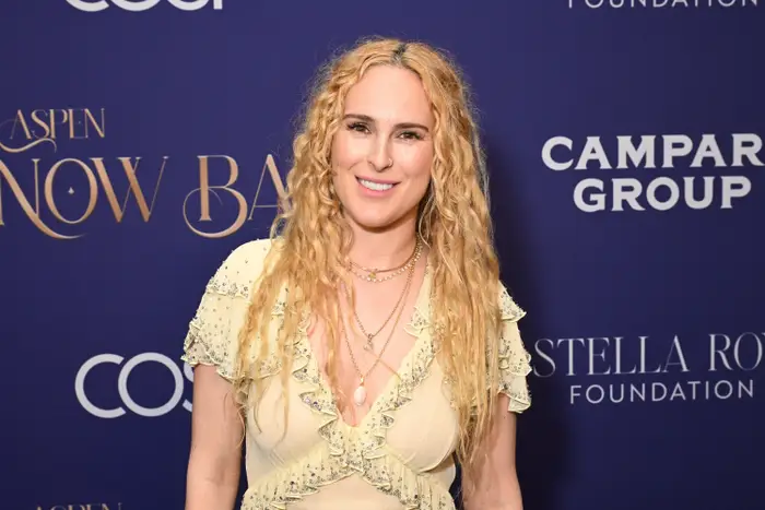 Person på rød løper i elegant, rynket kjole med lagdelte halskjeder, smilende mot bakteppet med eventlogoer