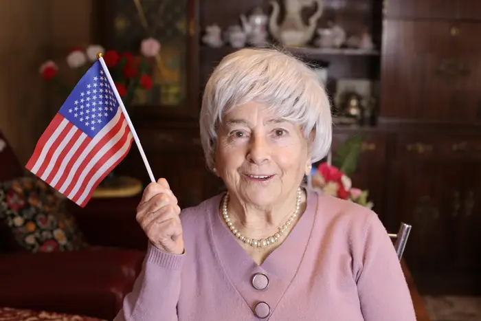 En eldre kvinne smiler og holder et lite amerikansk flagg. Hun har på seg et perlekjede og en cardigan med knapp, sittende i et koselig rom