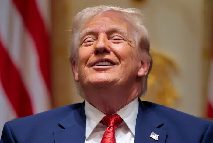 En person i dress og rødt slips smiler med lukkede øyne og virker avslappet og fornøyd. Det amerikanske flagget er uskarpt i bakgrunnen