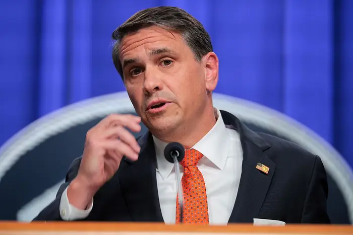 En mann i dress og oransje slips snakker ved et podium under en pressekonferanse