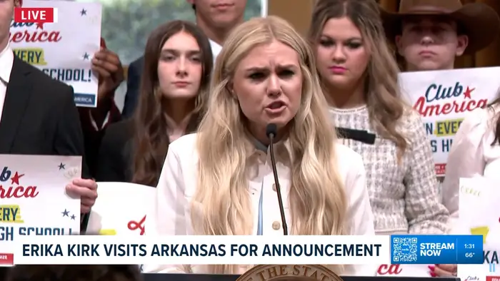 En kvinne holder en tale på et podium omgitt av folk som holder skilt, muligens på et skolerelatert arrangement. På banneret står det: "ERICA KIRK BESØKER ARKANSAS FOR KUNNGJØRING."