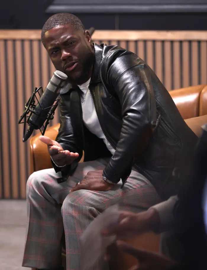 Person i skinnjakke og rutete bukser snakker inn i en mikrofon og gestikulerer med én hånd mens han sitter i studio.