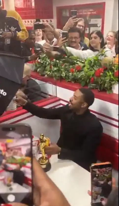 En person i formell antrekk sitter ved et bord med en pris. Folk i bakgrunnen tar bilder og fanger det festlige øyeblikket på et uformelt sted