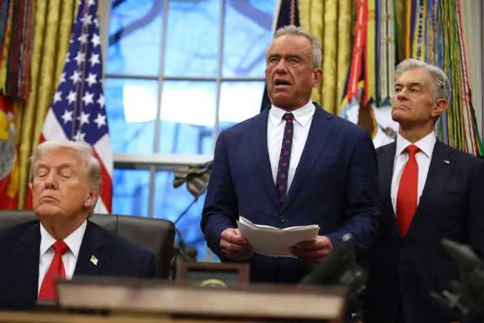 Tre menn i dress står i en formell kontorsetting, med flagg i bakgrunnen. En av dem holder et dokument og ser ut til å snakke