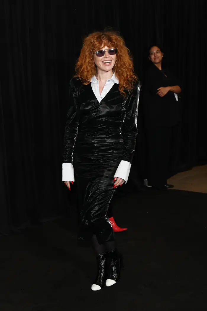 Natasha Lyonne med krøllete hår iført en elegant, langermet kjole og solbriller, smilende på et teppe