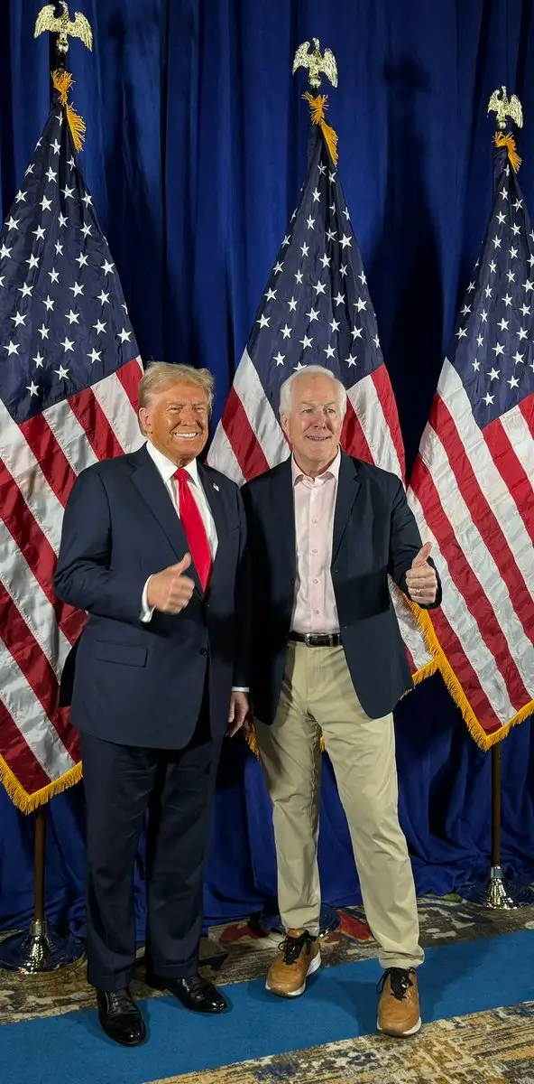 To menn står og smiler med tommel opp foran flagg. Mannen til venstre har på seg dress og rødt slips, den andre i blazer og kaki.