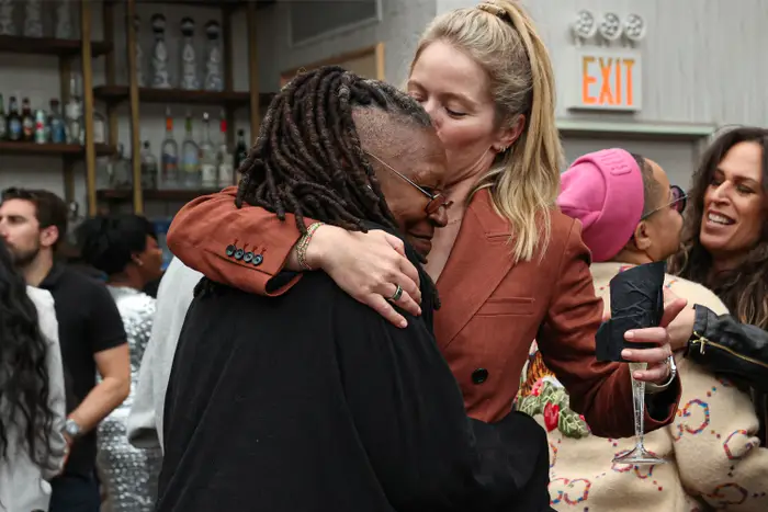 To personer omfavner seg varmt på en sosial begivenhet. Den ene bruker blazer, den andre har dreadlocks. Folk blander seg i bakgrunnen