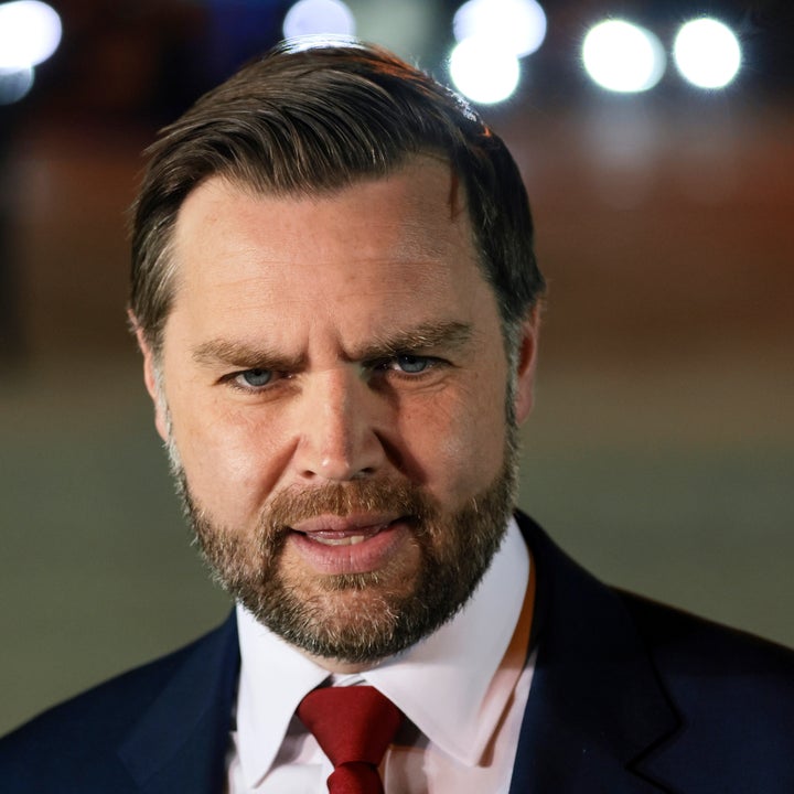 Mann med pent stylet hår og skjegg i dress og slips, stående utendørs om natten med lys bak seg