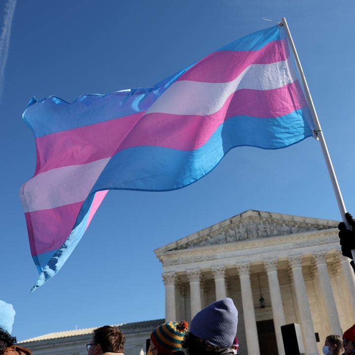 Person som holder et transgender pride-flagg under et rally foran en stor bygning med søyler