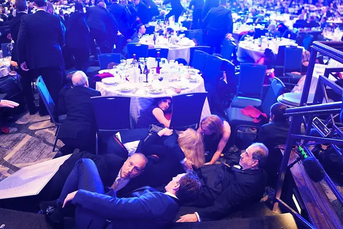 Folk i formell antrekk ligger på bakken under bord ved en formell begivenhet, og ser ut til å ta dekning eller delta i en leken aktivitet