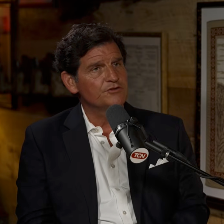 En mann i mørk dress snakker inn i en mikrofon på "The Tucker Carlson Show"-settet, med et innrammet bilde i bakgrunnen
