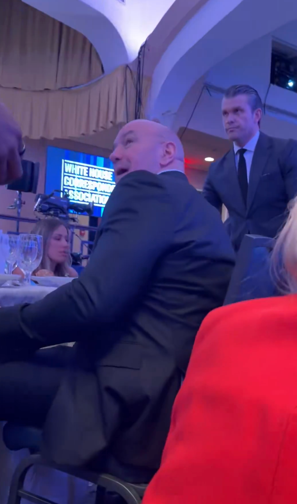 En mann i dress sitter på et formelt arrangement med bord og deltakere synlige i bakgrunnen
