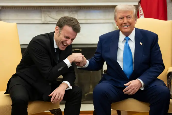 To menn i dress sitter i stoler, håndhilser og smiler. Man lener seg frem og ler. De ser ut til å være i en formell setting