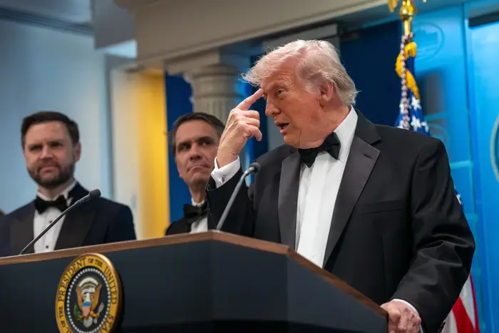 To menn står bak et podium med en presidentsegl mens en tredje mann i smoking gester
