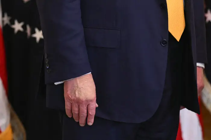 Donald Trump, med sminke på hånden, står i en formell dress med et oransje slips foran et amerikansk flagg i bakgrunnen