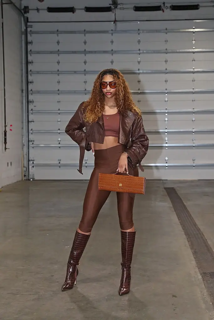 Personen poserer selvsikkert i et stilig antrekk med en crop top, skinnjakke, gjennomsiktige leggings, støvler og solbriller, og holder en matchende clutch