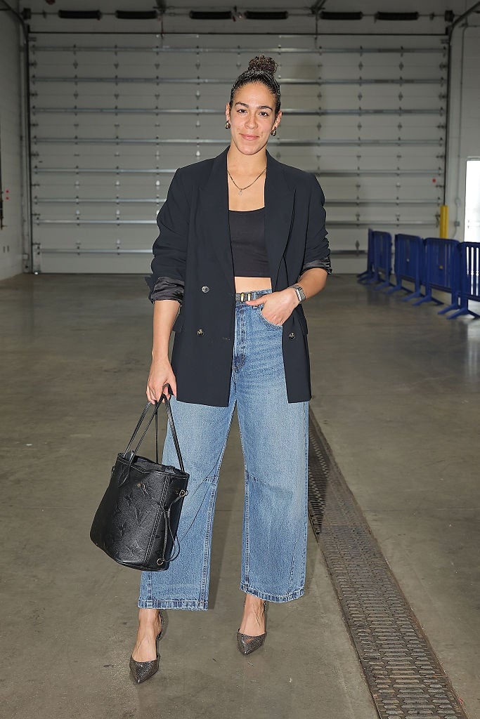 Person i et casual-chic antrekk med blazer, cropped topp og jeans, holder en stor veske, stående innendørs