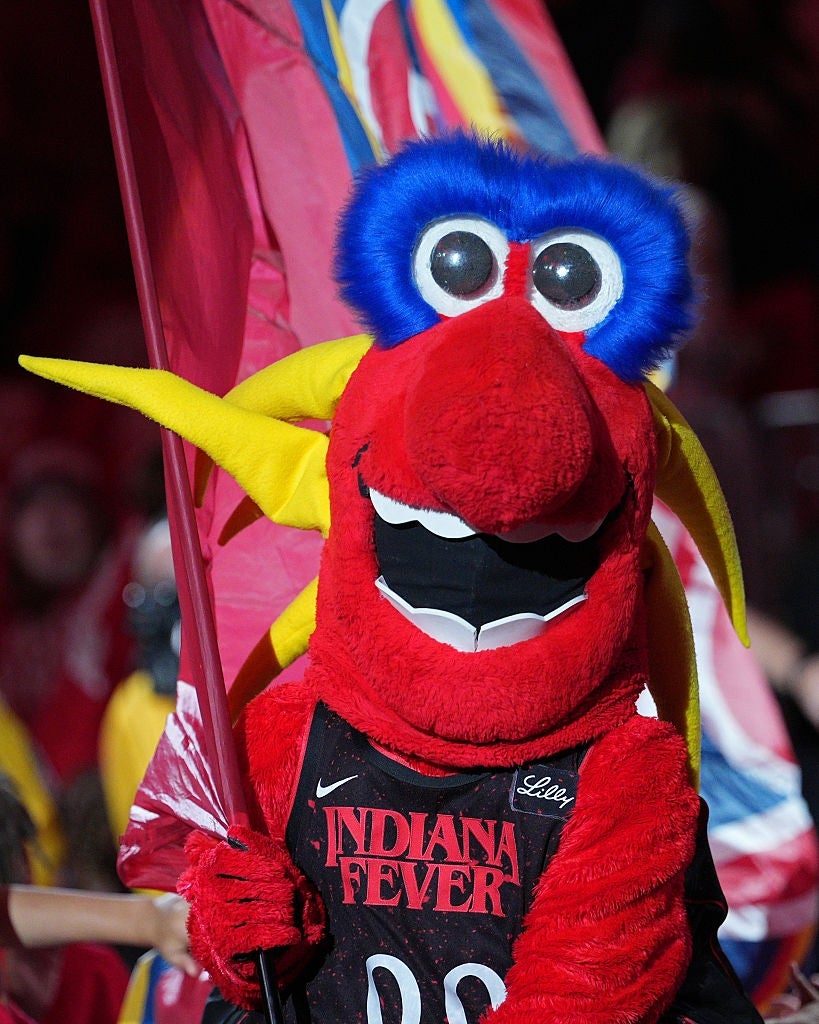 En rød og gul maskot med et stort fjærhode holder et flagg på et sportsarrangement, iført en Indiana Fever-trøye