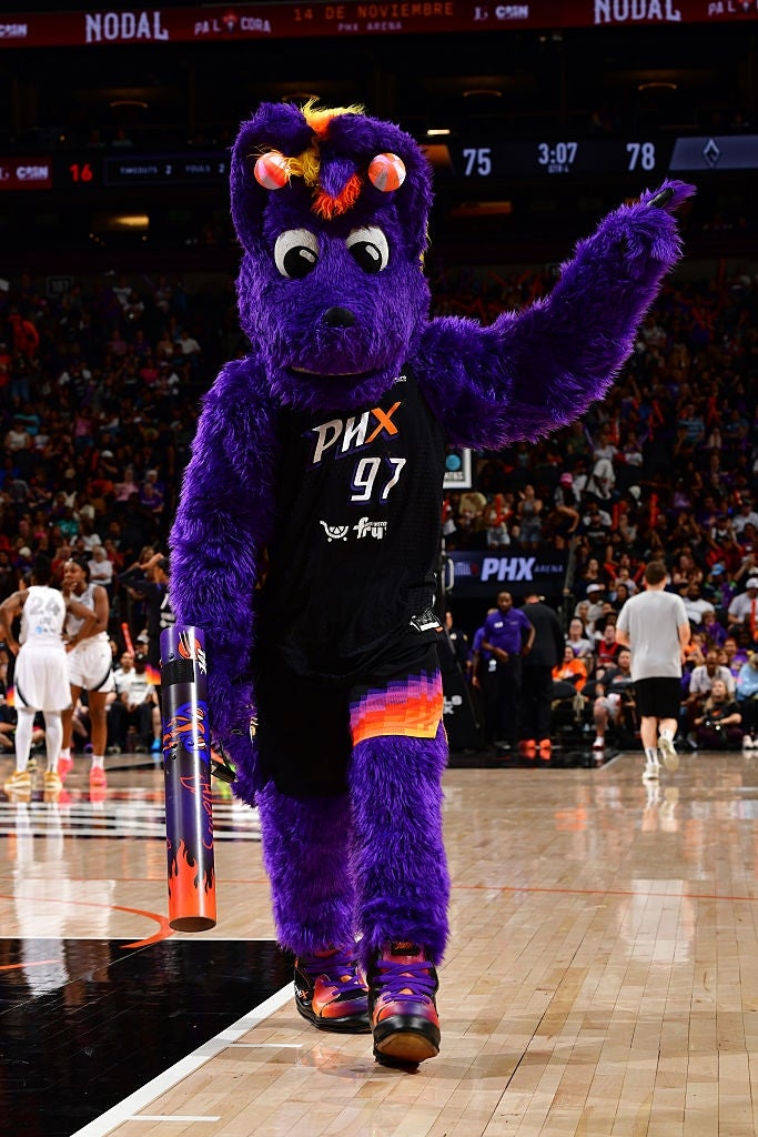 Maskot i lilla lodne antrekk med basketballuniform, som underholder et publikum under en basketballkamp