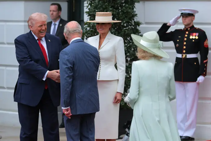 To menn i formelle dresser hilser på hverandre utendørs, med to kvinner i elegante hatter i nærheten, og en marinehilsen i bakgrunnen