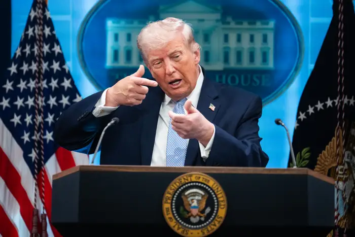 En person snakker ved et podium med en presidentsegl, gestikulerer med begge hender, med amerikanske flagg og et hvitt hus som bakteppe synlig