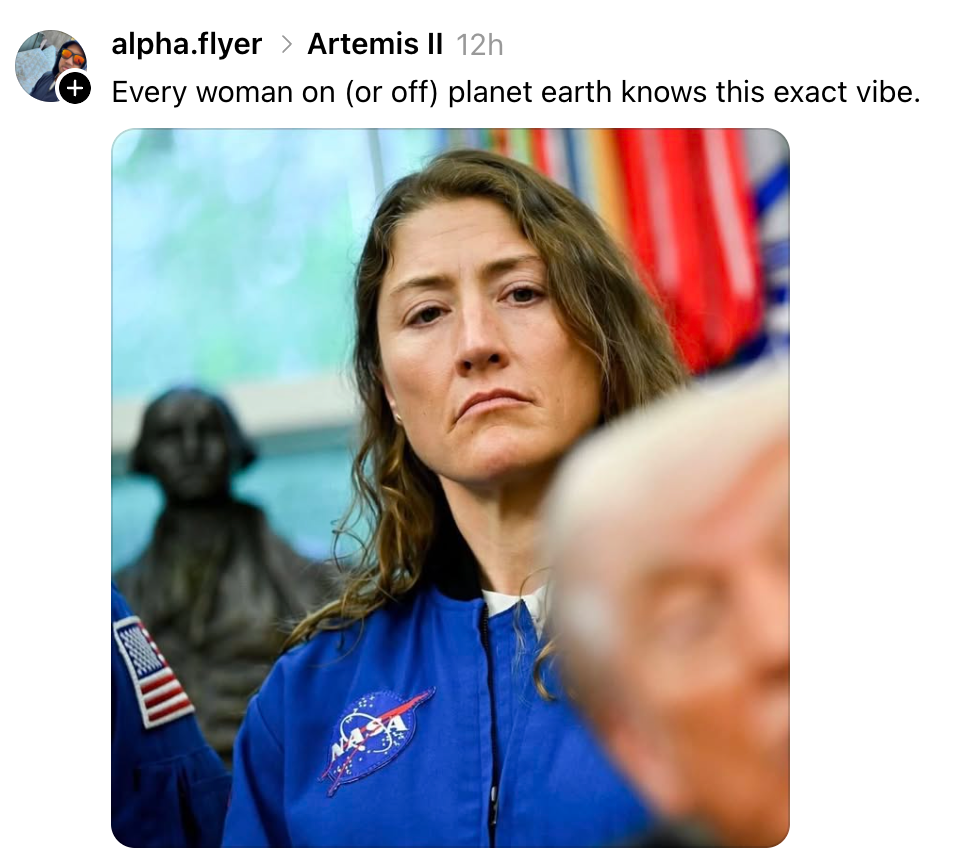 En astronaut i en NASA jumpsuit ser alvorlig ut, med en gruppe mennesker og flagg i bakgrunnen